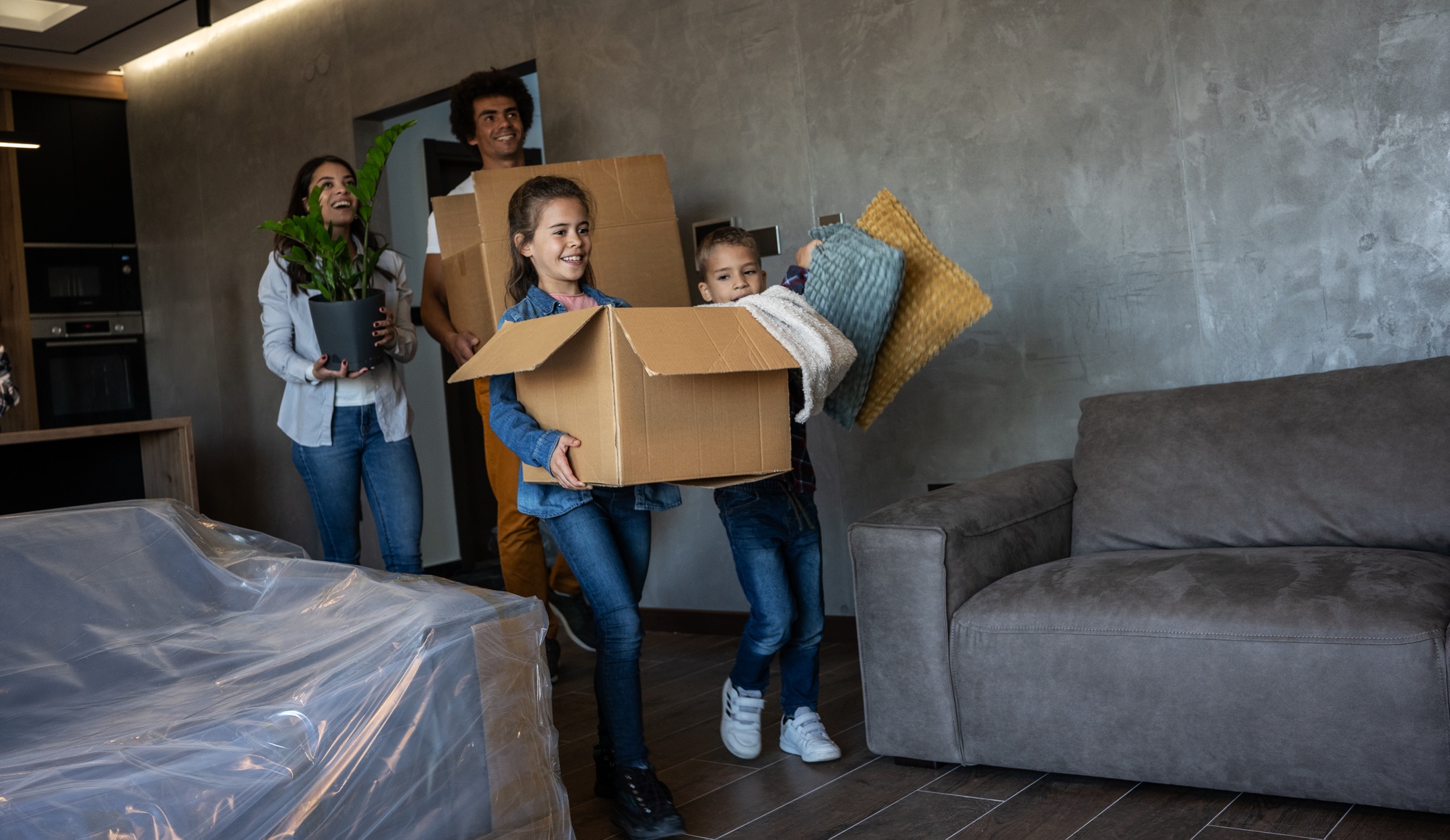 Famille transportant des cartons lourds et des objets dans un salon moderne, prêt à déménager, avec une plante et canapé visibles. check-list déménagement à imprimer intégré naturellement.