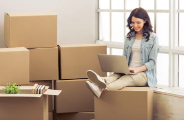 Jeune femme assise sur des cajas avec un ordinateur portable, entourée de cartons de déménagement, dans une pièce lumineuse. Coût moyen d'un déménagement de longue distance.